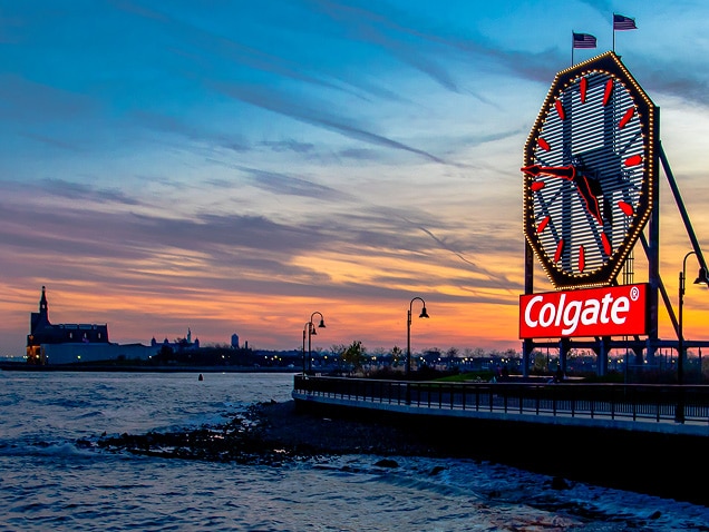 Kystlinje med Colgate-logo i baggrunden under et stort ur med røde visere.